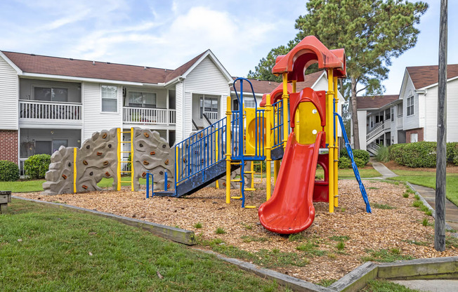 Outdoor playground with slide and climbing wall with wood chips on the ground surrounded by apartment buildings.