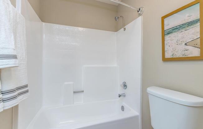 A clean and modern bathroom featuring a white bathtub with a showerhead, a white toilet, and a small framed image of a beach on the wall. Soft towels hang nearby, adding to the tidy and inviting atmosphere.