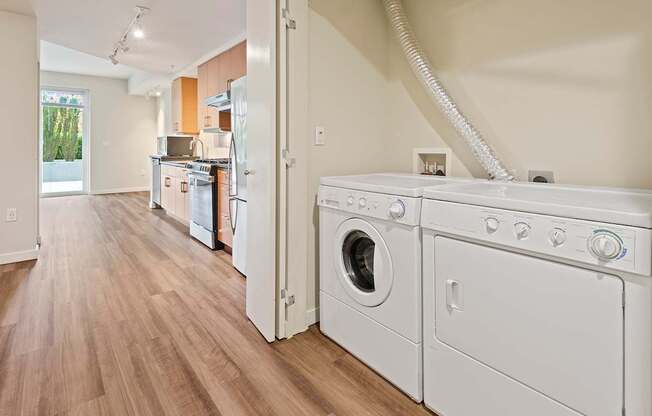 A white washer and dryer are in a laundry room with a kitchen in the background.