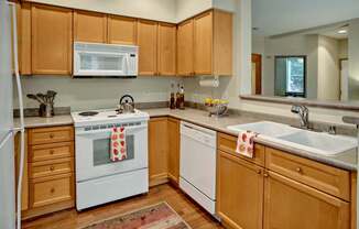 A welcoming kitchen here at Portsmith showcasing light wood cabinetry, neutral countertops, a double-basin sink, a dishwasher, and a built-in microwave above the range, complemented by warm wood-style flooring, simple countertop décor, and an open pass-through that connects the kitchen to the adjacent living space.