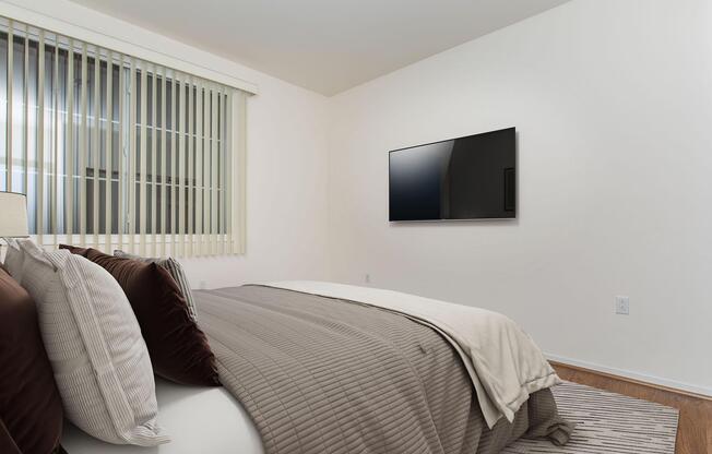 A simple, modern bedroom featuring a neatly made bed with gray and brown bedding. There are two pillows on the bed and a lamp on a nightstand beside it. A large flat-screen TV is mounted on the wall opposite the bed, and the room has light-colored walls and natural light streaming through the window with blinds.