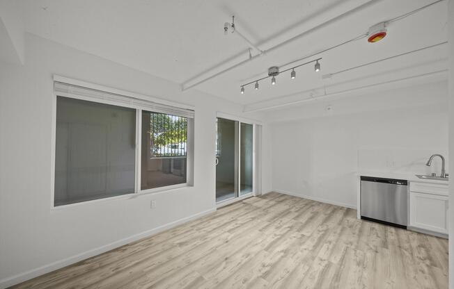 A bright, empty living space with white walls, large windows, and sliding glass doors that open to a patio. The floor is light-colored wood, and there is a kitchen area with a dishwasher and sink in the corner. The ceiling features exposed ductwork and track lighting.