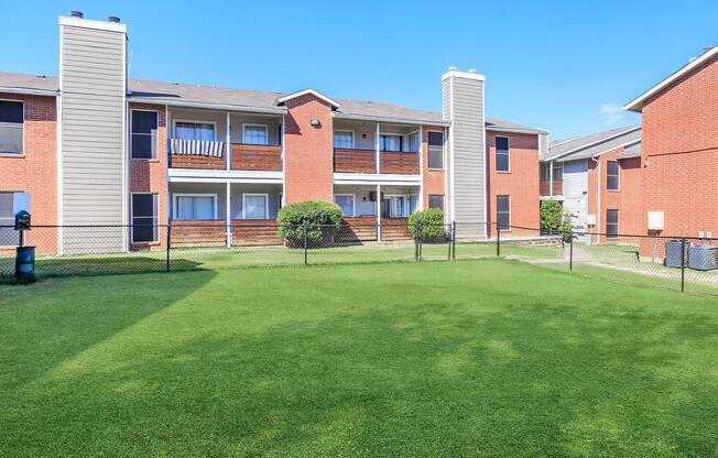 A well-maintained apartment complex featuring red brick buildings with balconies, surrounded by a green lawn. The scene includes a fenced area, clear blue sky, and well-kept landscaping, creating a welcoming atmosphere for residents.