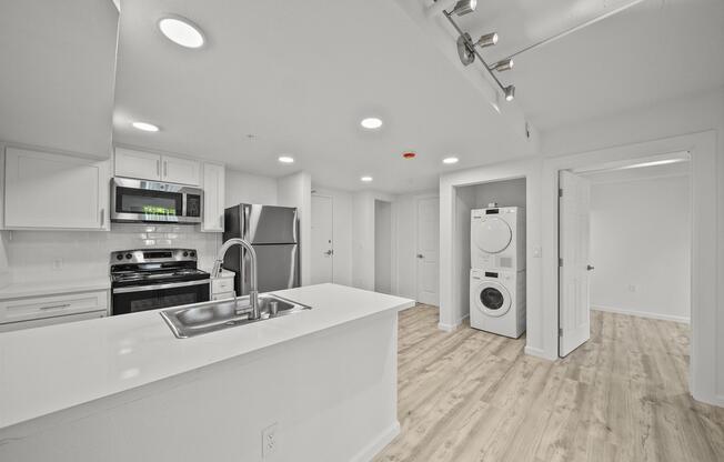Modern kitchen and laundry area with stainless steel appliances, including a stove and refrigerator. The open layout features a white countertop and cabinetry, with a washer and dryer in a dedicated space. The flooring is light wood, and the walls are painted white, creating a bright and airy atmosphere.