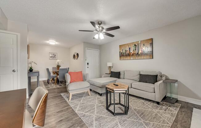 A cozy living room featuring a light gray sectional sofa with accent pillows, a wooden coffee table, and a decorative rug. Thereâs a ceiling fan, a small side table with a lamp, and a dining area in the background with a round table and chairs. The walls are decorated with modern art.
