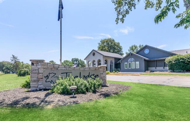A well-manicured lawn in front of a building with a modern design and large windows. A stone sign with the name "Tuleadise" is prominently displayed, alongside a flagpole. Trees and greenery are visible in the background under a clear blue sky, creating a welcoming outdoor atmosphere.