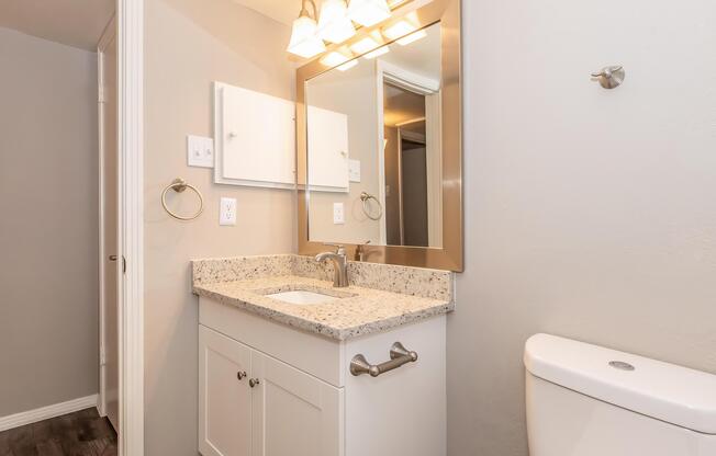 A modern bathroom featuring a white vanity with a granite countertop, a mirror above the sink, and a wall-mounted towel holder. A white toilet is visible on the right side, with neutral-colored walls and a light fixture above the mirror. The flooring is dark wood.