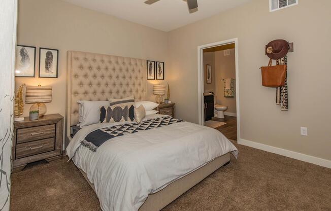 A cozy, modern bedroom featuring a tufted beige headboard with decorative pillows. Two bedside tables with lamps are on either side of the bed. Art prints adorn the wall above the bed. A light tan color scheme and plush carpet create a warm atmosphere. A bathroom is visible through an open door in the background.