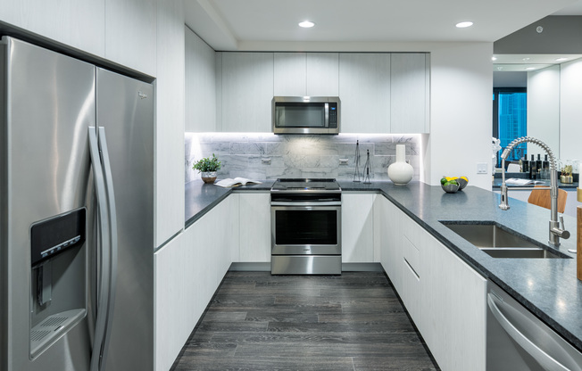 Kitchen with dark wood-style floors, stainless steel appliances, white frameless and handleless cabinets, gray quartz countertops, and an undermount sink with a detachable faucet.