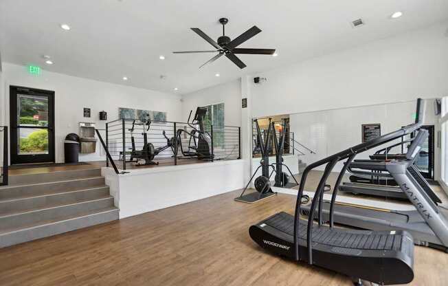 A gym with a treadmills, equipment, a staircase, and a ceiling fan.