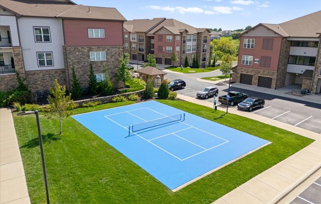 A tennis court is located in a grassy area in front of apartment buildings.