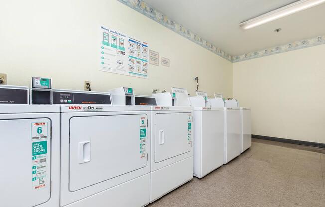 A laundry room containing several white washing machines and dryers lined up against a wall. The room has a light-colored, clean appearance with a bulletin board and signs on the wall providing laundry instructions. The floor is tiled, and there is overhead lighting.