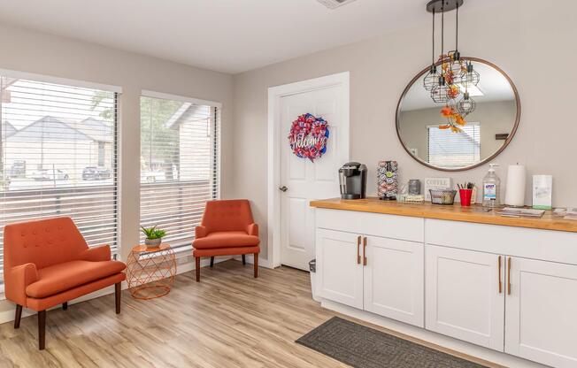 Cozy waiting area featuring two orange chairs, a circular mirror, and a wooden counter with coffee supplies. A red and blue wreath adorns the wall, while natural light streams in through the windows with blinds. A small plant and various brochures are visible on the counter.