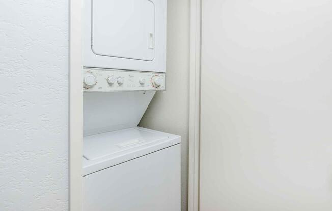 A stacked washer and dryer unit in a narrow laundry closet. The appliances are white and feature control knobs. The surrounding walls are light-colored, and there's a door partially visible on the right side. The overall setting is clean and functional.
