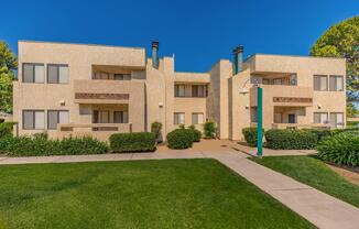 Modern multi-unit apartment building with a light beige exterior, featuring balconies and green landscaping. A well-maintained pathway leads to the entrance, with manicured lawns and shrubs surrounding the property. Clear blue sky serves as a backdrop to the structured design of the building.