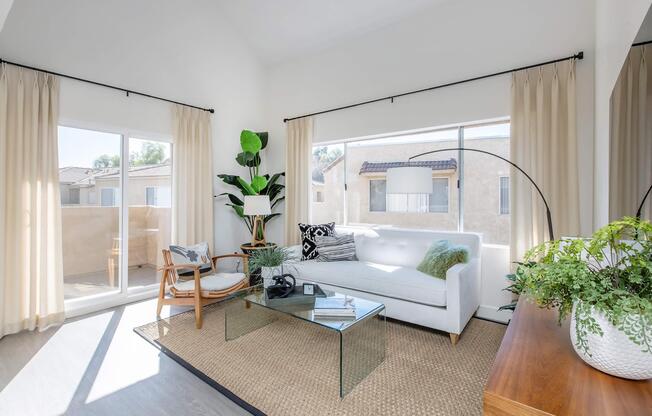 A bright and airy living room featuring a white sofa with decorative pillows, a glass coffee table, and a woven rug. Large windows with sheer curtains allow natural light to flood the space. There are indoor plants and a wooden side table, creating a cozy, modern atmosphere.