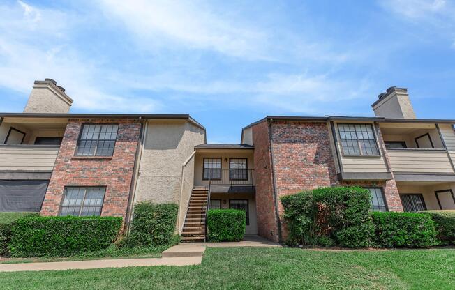 Two-story apartment building with a mix of brick and beige siding. The building features two sets of stairs leading to upper units, surrounded by green shrubs and a grassy area. A clear blue sky is visible above, creating a bright and inviting atmosphere.