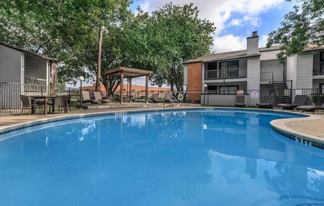 A clear blue swimming pool surrounded by lounge chairs and shaded areas, with trees and buildings in the background.