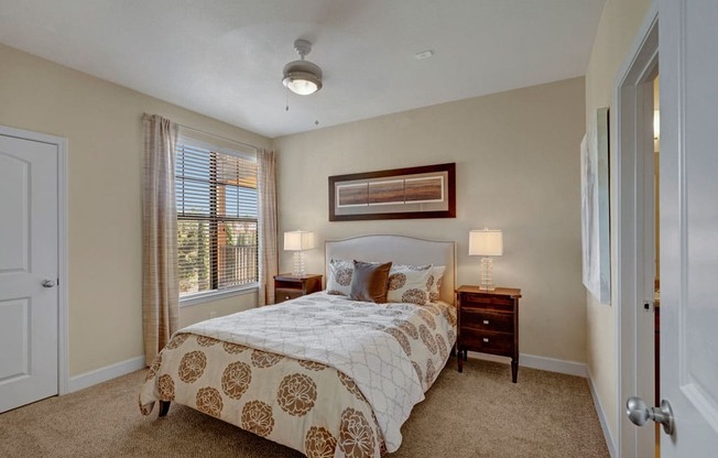 A spacious bedroom here at Tuscany at Faudree with plush carpet, soft neutral walls, and a comfortable bed centered between matching nightstands and lamps. A large window with blinds and curtains brings in natural light, while framed wall art and a ceiling fan add a calm, inviting feel to the room.