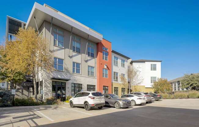 A parking lot with cars and a building with a white car parked in front at Infinity on the Point Apartments, Dallas, TX