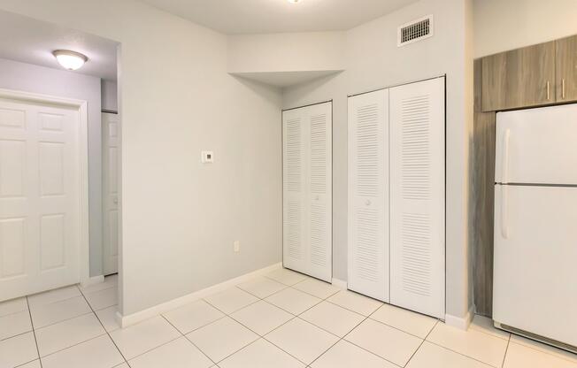 An interior space featuring light gray walls, tiled flooring, and a large white refrigerator. There are two white sliding door closets on the right, and a doorway to the left leading to another room. The ceiling has a light fixture, creating a bright and open atmosphere.