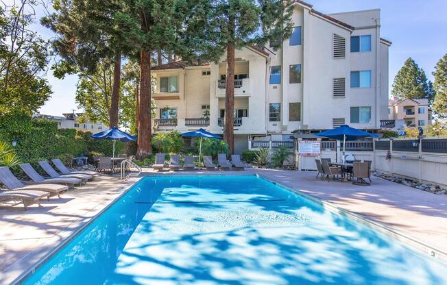 A clear blue swimming pool surrounded by lounge chairs and umbrellas, with tall trees in the background. A three-story residential building is visible behind the pool, featuring large windows and balconies. The area is well-kept, suggesting a relaxing outdoor space.