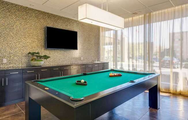 A pool table with a green cloth and white balls is in the foreground of a room with a television and patterned wallpaper.