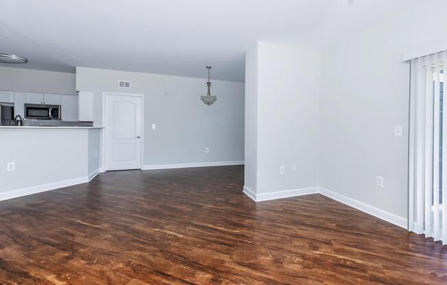 A spacious, empty living area featuring hardwood flooring, light-colored walls, and a single light fixture hanging from the ceiling. To the left, there's a view of a kitchen area with modern appliances visible. Large sliding glass doors open to a balcony, allowing natural light to fill the room.