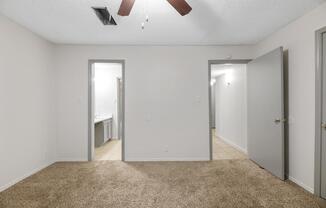 A spacious room with light beige carpet and plain white walls. There are two doorways on the left leading to other rooms, and a partially visible bathroom on the far left. The ceiling features a ceiling fan with wooden blades. Soft lighting creates a bright and inviting atmosphere.