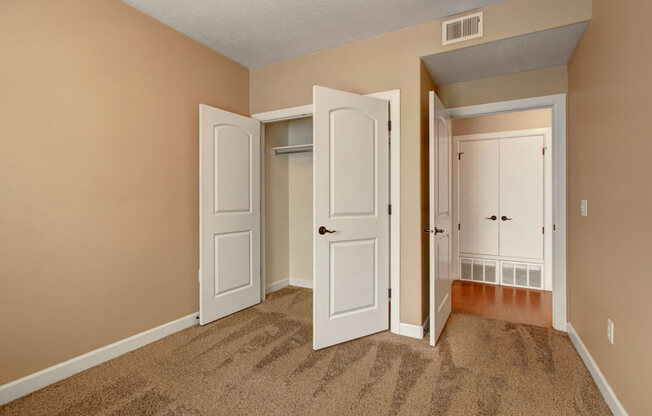 A bedroom here at Bridges with tan walls, soft brown carpet, an open white double-door closet, and a hallway view toward matching white doors and warm wood flooring.