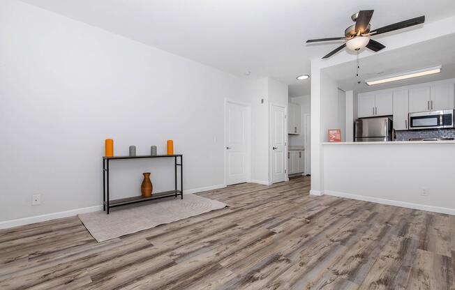 A modern, minimally furnished living space featuring light wood flooring. A small black table with decorative vases in orange and gray colors is positioned against a white wall. The background shows an open kitchen area with stainless steel appliances and white cabinetry. Soft beige rug on the floor adds warmth to the room.