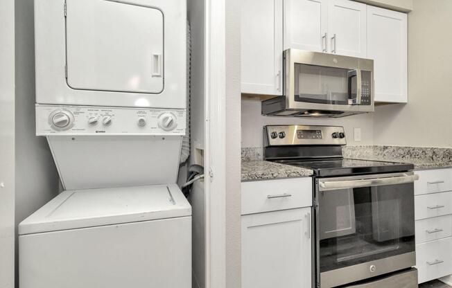 A laundry area featuring a stacked washer and dryer is shown on the left, while the right side displays a modern kitchen with white cabinetry, a granite countertop, a microwave, and an oven.