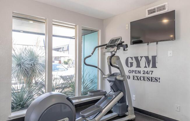 A modern gym space featuring an elliptical machine, a wall-mounted television, and large windows with natural light. The wall displays text that reads "GYM OPEN 24/7 NO EXCUSES!" with a minimalist design and potted plants visible outside.