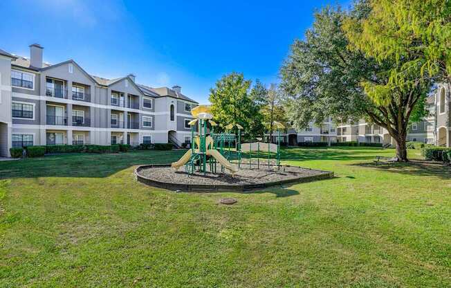 Spacious outdoor playground area at Saxony at Chase Oaks Apartments, featuring a colorful play structure with slides and climbing elements, surrounded by lush green lawns and mature trees. The modern apartment buildings in the background showcase elegant gray and white exteriors with balconies.