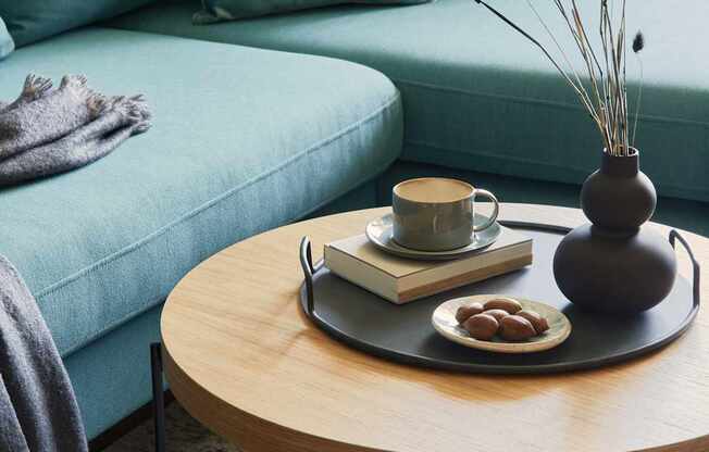 A living room with a blue couch, a wooden table with a vase, a book, a cup, and some fruit.