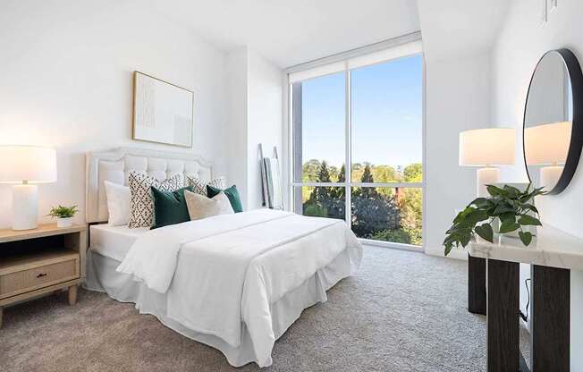 A bedroom with a large bed, a nightstand, a lamp, a mirror, and a window with a view of trees.