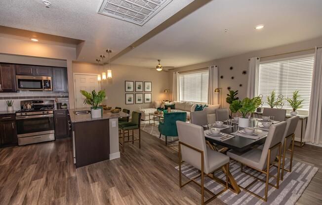 A modern open-concept living space featuring a stylish kitchen with stainless steel appliances, a dining area with a large table set for six, and a cozy living room with green accent chairs. Natural light streams in through large windows, and decorative plants add a touch of greenery to the space.