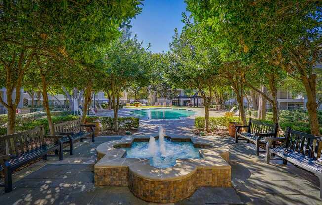 A shaded courtyard at Saxony at Chase Oaks Apartments in Dallas, TX, featuring a decorative fountain, benches, and lush greenery, with views of the pool area in the distance.