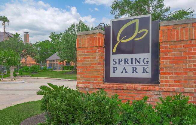 Sign for Spring Park featuring a green leaf logo, surrounded by landscaped greenery. The area has a well-maintained pathway and residential buildings visible in the background under a partly cloudy sky.