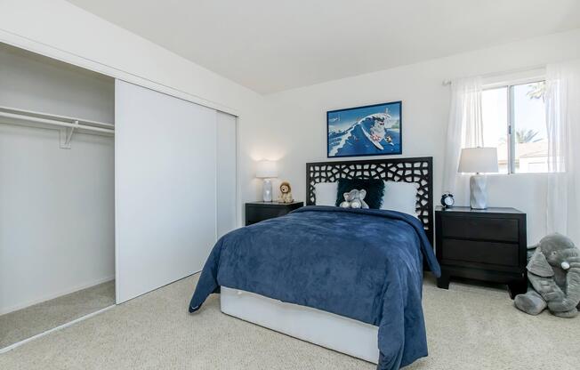 A cozy bedroom featuring a twin bed with a blue blanket, a nightstand with a lamp, and a wall-mounted artwork of a surfing scene. There's a closet with sliding doors to the left and large windows allowing natural light to fill the room. Soft beige carpet flooring completes the inviting atmosphere.