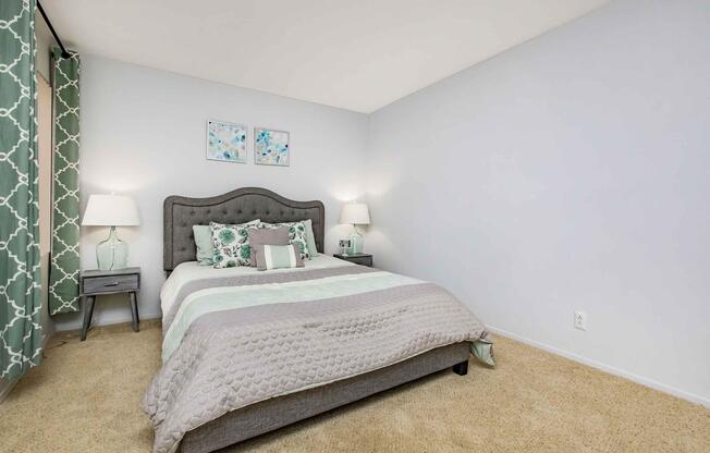 A neatly arranged bedroom featuring a gray upholstered bed with a decorative quilt and pillows. Two bedside lamps sit on matching nightstands. The walls are painted light gray, and there are two framed artworks above the bed. Green patterned curtains complement the decor, with plush carpeting covering the floor.