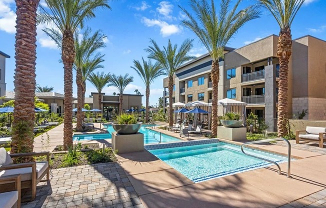 A resort-style pool courtyard here at Reflect at Dobson Ranch featuring a rectangular swimming pool with built-in steps and handrails, surrounded by tall palm trees, modern apartment buildings, lounge seating, shaded umbrellas, and landscaped walkways that create a relaxed and inviting outdoor community space.