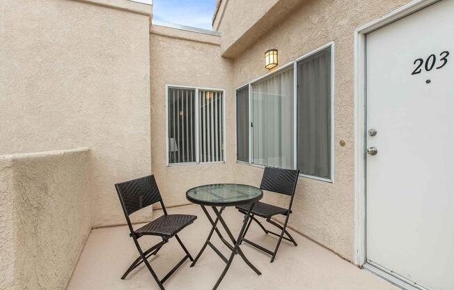 A small patio area featuring a round glass table with two black folding chairs. The space is surrounded by beige exterior walls and has a door marked "203." There are two windows with white vertical blinds allowing natural light into the area.