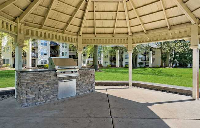 A gazebo with a stone pillar and a metal door is surrounded by a concrete floor and a wooden roof.