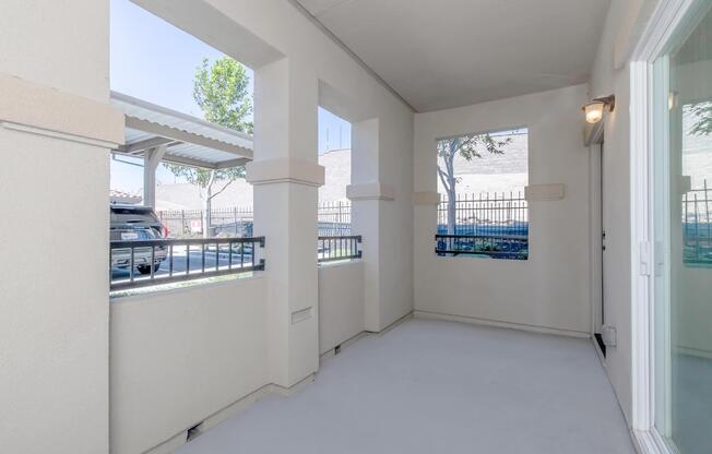 A small, covered outdoor space with light-colored walls and a smooth floor. Two openings provide views outside, featuring a parked car and a fenced area with trees in the background. The space is well-lit with a light fixture on the wall.