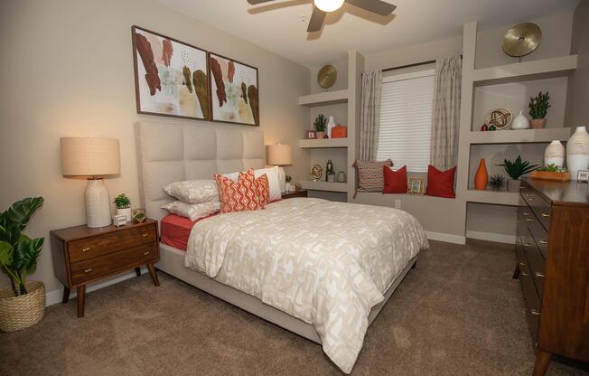 A cozy bedroom featuring a light-colored bed with a patterned duvet, flanked by two bedside lamps. The walls are adorned with abstract art, and there are decorative shelves with plants and accessories. Natural light streams in through a window with patterned curtains, adding warmth to the inviting space.