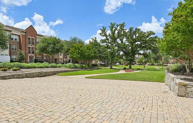 A brick pathway leads to a grassy area with trees and a building in the background.
