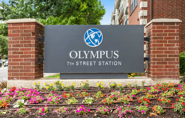 A sign for Olympus 7th Street Station is framed by brick pillars and adorned with colorful flowers.
