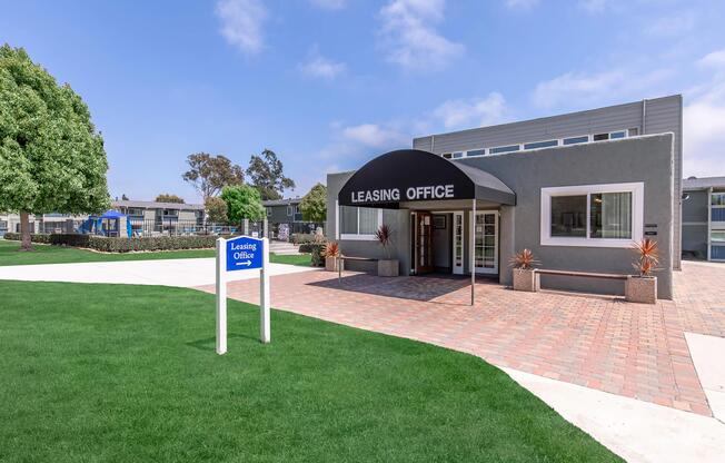 Leasing office building with a black awning and sign, surrounded by green grass and trees. A blue sign with "Leasing Office" is visible in front. The entrance features large windows, and the area includes a paved walkway leading to the building. Clear blue sky in the background.