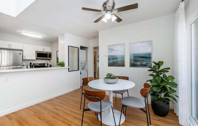 A bright and modern dining area featuring a round white table surrounded by four chairs, a green potted plant in the corner, and two framed ocean-themed pictures on the wall. The space has wood flooring and is illuminated by a ceiling fan light. A kitchen area is visible in the background.